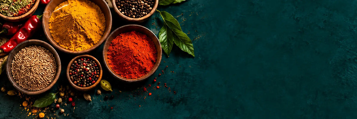 An overhead flat-lay of various colorful ground spices in small brown bowls, including turmeric, red chili powder, cumin, and peppercorns, arranged on a dark teal textured background with scattered whole spices and green leaves.